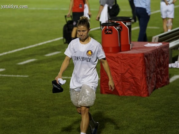 A person wearing a white sports jersey with 'CONSERVICE' written on it and ice packs wrapped around both knees is walking on a grass field. The background includes a red table with large cooler jugs on it and several other people, some blurred, standing around. The person appears to be holding a black cap and a cloth.