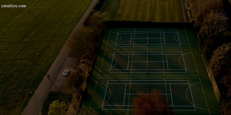 An aerial view of multiple tennis or sports courts next to a green recreational field, bordered by trees. The courts are marked with lines and look well-maintained. Shadows cast by trees stretch over parts of the courts and pathways.