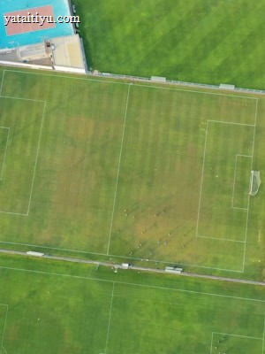 An aerial view of a sports complex featuring multiple green grass fields, including football pitches with marked goalposts. Adjacent to the fields is a blue-roofed building and a smaller area with a tennis or basketball court. Surrounding the complex, there's a road lined with trees and a dirt area with sparse vegetation.