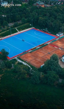 An aerial view of a large sports complex with a vibrant blue field surrounded by a red boundary and adjacent clay courts. The complex is nestled amidst dense greenery and trees, with a few buildings visible in the background. The setting appears to be serene and well-maintained, with a sense of spaciousness.
